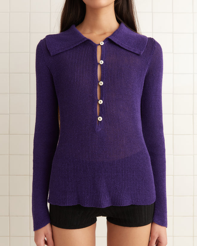 A person in a 1970s-inspired Quincy Top - Purple with pearl buttons and black shorts stands against a tiled wall.