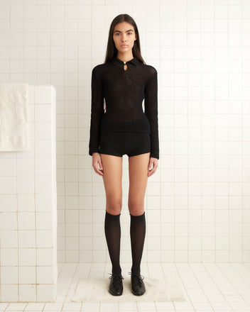 A person stands in a tiled room, wearing the Willows Pullover - Black, black shorts, and knee-high socks with black shoes. A towel hangs on the left wall.