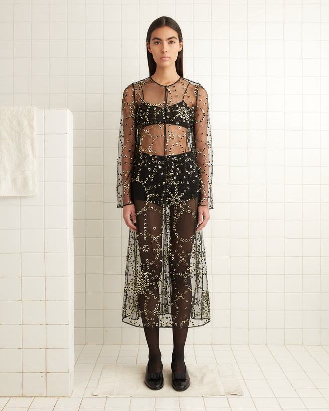 In a tiled room with white walls and minimalist decor, a person stands in a translucent, beaded dress, enhanced by the shimmer of the **Sheer Fête Duster** under the soft lighting.