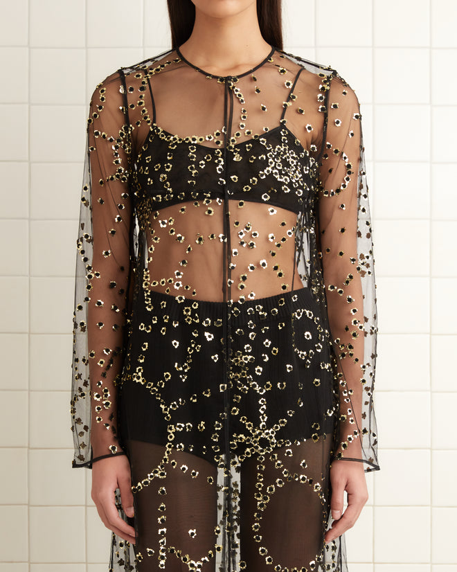 In a tiled room with white walls and minimalist decor, a person stands in a translucent, beaded dress, enhanced by the shimmer of the **Sheer Fête Duster** under the soft lighting.