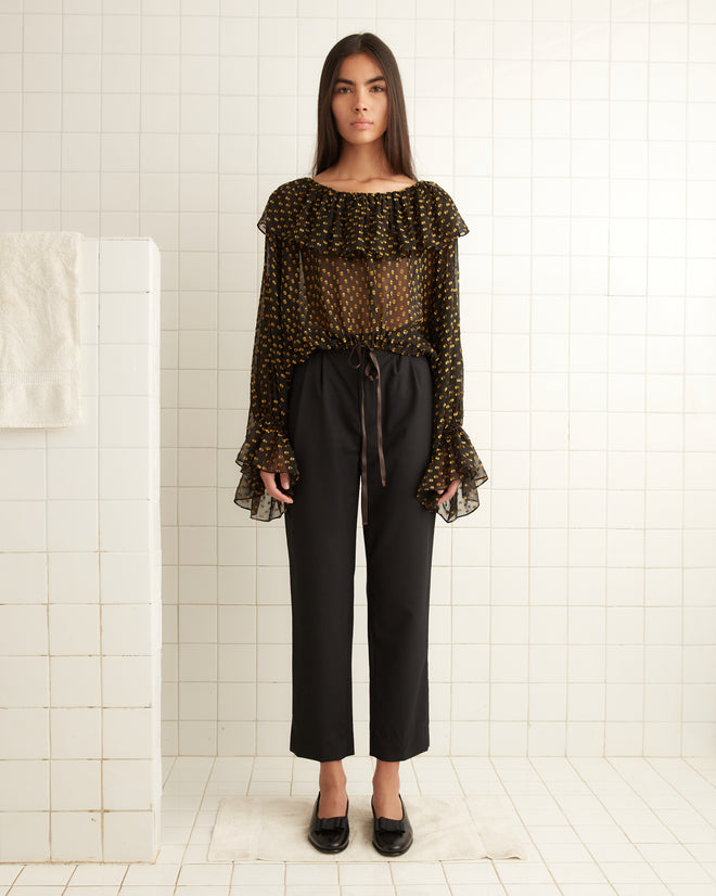 A woman in a Stadium Dot Blouse featuring sheer fabric with a ruffled collar and cuffs, paired with black pants and shoes, stands gracefully in a tiled room.