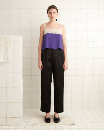 A person stands in a tiled room wearing the Iris Lace Tank, a purple top crafted from exquisite hand-embroidered fabric, and black pants, with hands relaxed by their sides.