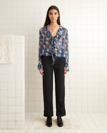 A woman in a Daisy Blouse with a 1930s floral print and black pants stands gracefully in a tiled room, with a towel hanging on a nearby divider.