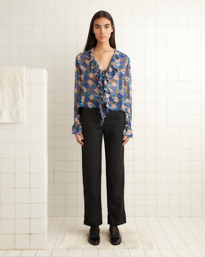 A woman wearing the Daisy Blouse, featuring sheer crepe fabric with floral patterns and ruffles, along with black pants stands in a tiled room.