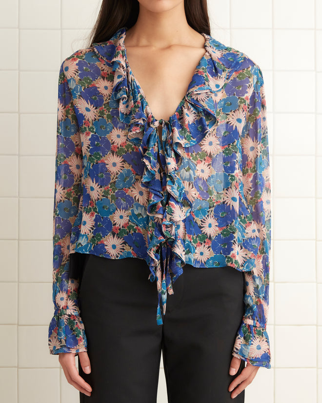 A woman wearing the Daisy Blouse, featuring sheer crepe fabric with floral patterns and ruffles, along with black pants stands in a tiled room.