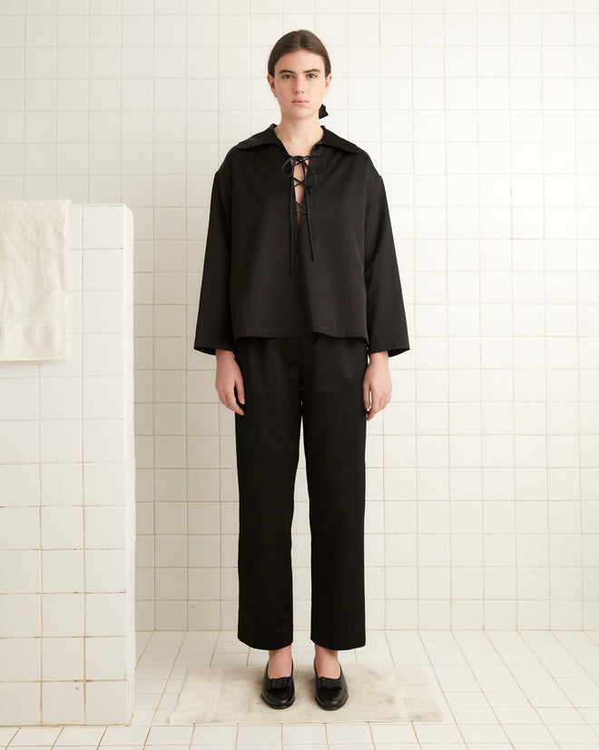 A person wearing the Sheen Bonnie Shirt in black stands in a bathroom with white tiled walls and a towel hanging nearby, their ensemble subtly enhanced by the satin weave texture of the shirt.