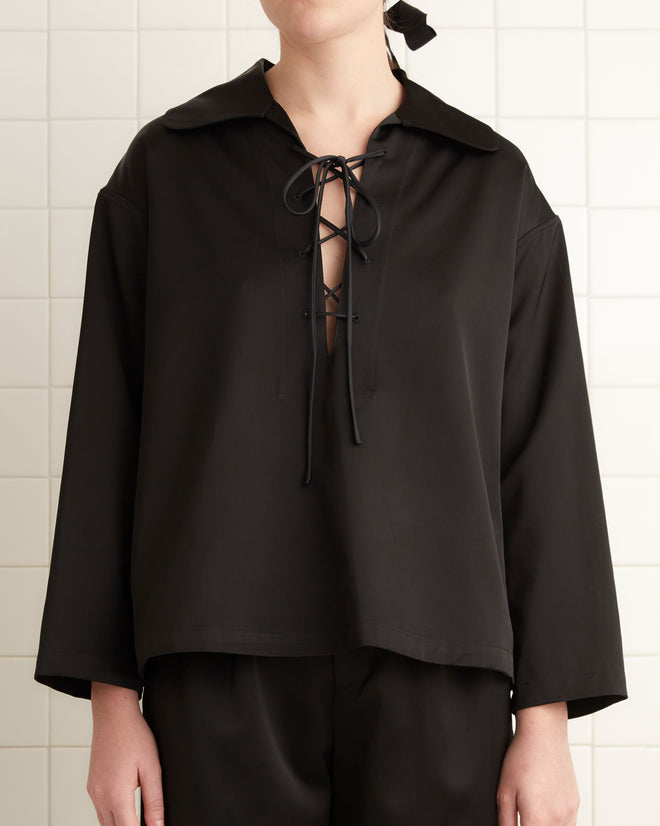 A person wearing the Sheen Bonnie Shirt in black stands in a bathroom with white tiled walls and a towel hanging nearby, their ensemble subtly enhanced by the satin weave texture of the shirt.