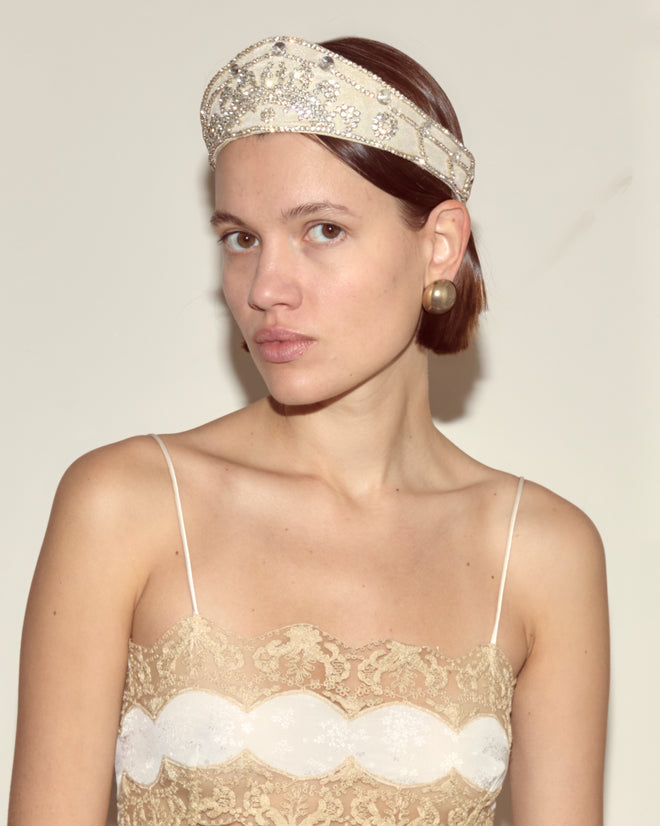 A woman wearing the Willow Crown - White and large gold earrings gazes at the camera in a lace dress, channeling the elegance of a 1930s silhouette.