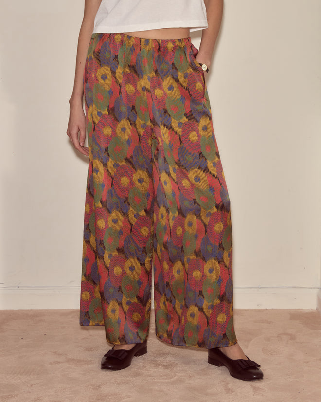 A person wearing Fruit Pit Pants—wide-leg, colorful floral silk seersucker bottoms—with a white top and dark shoes stands on a beige carpet.