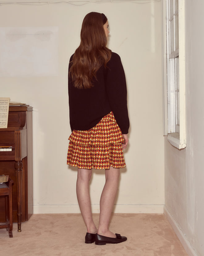 A woman with long brown hair stands indoors, facing a wall, wearing a black top and the Orana Ruffle Skirt inspired by a 1960s French pattern.