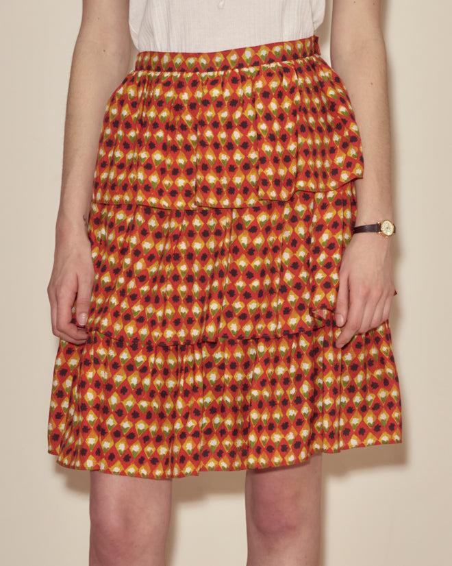 Person wearing the Orana Ruffle Skirt in scarlet silk, featuring three tiers, paired with a white top and black wristwatch, facing forward.