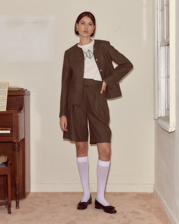 A woman stands indoors by a piano wearing Morris Houndstooth Shorts with D-ring waist adjusters, a plaid blazer, white shirt, knee-high socks, and black shoes.
