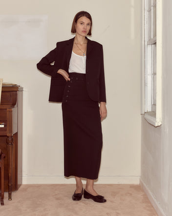 A woman in a black blazer and white top stands indoors by a piano and window, wearing the Riding Twill Sailor Skirt.