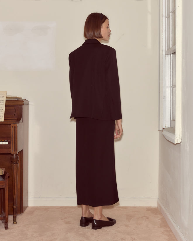 A woman in a Riding Twill Sailor Skirt stands indoors, facing away near a piano and window, radiating timeless elegance.