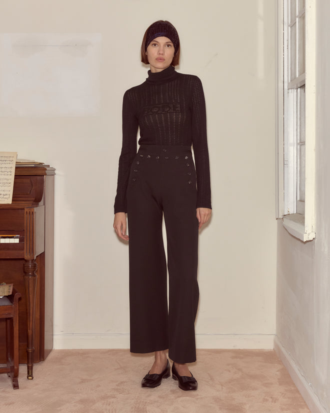 A woman in a black knit top, Riding Twill Sailor Trousers - Black, and a headband stands indoors by a piano and window.