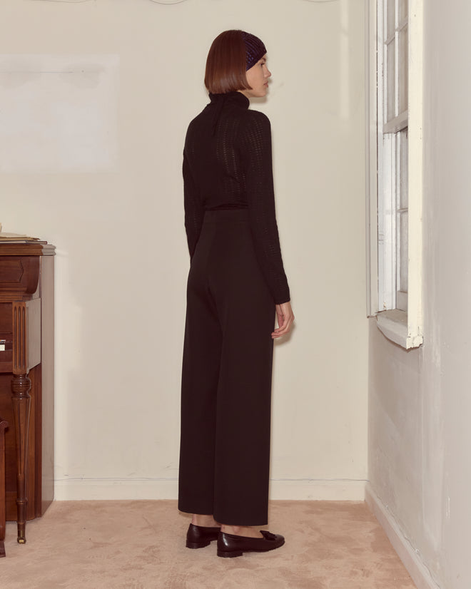 A woman in Riding Twill Sailor Trousers - Black and a black top stands sideways in a simple room near a window.