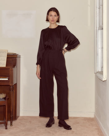 Woman wearing Astor Port Trousers and a black blouse stands indoors by a piano, near a window on beige carpet.