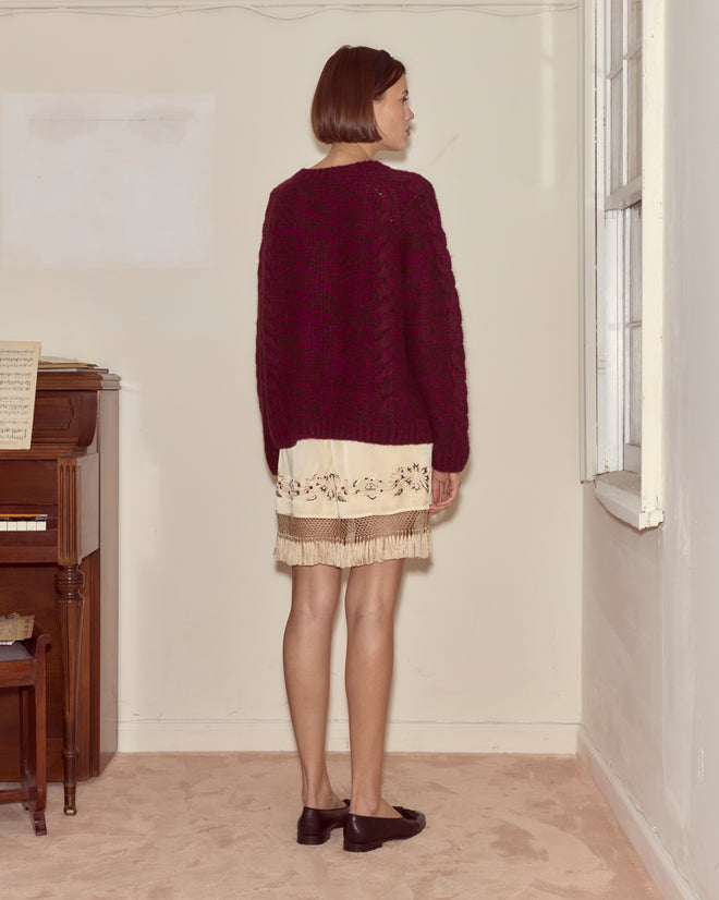 A woman in a burgundy sweater and the Phantasm Fringe Skirt stands indoors near a piano, facing a window.