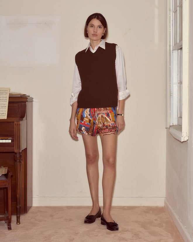 A woman stands by a piano in a softly lit room, wearing a white shirt, black vest, and Manhattan Island Shorts.