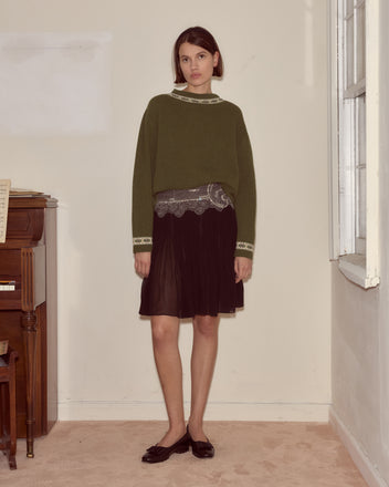 A woman wearing an olive green sweater, the Ensemble Skirt in black silk georgette with beading, and black flats stands indoors by a piano and window, evoking a 1930s reproduction style.