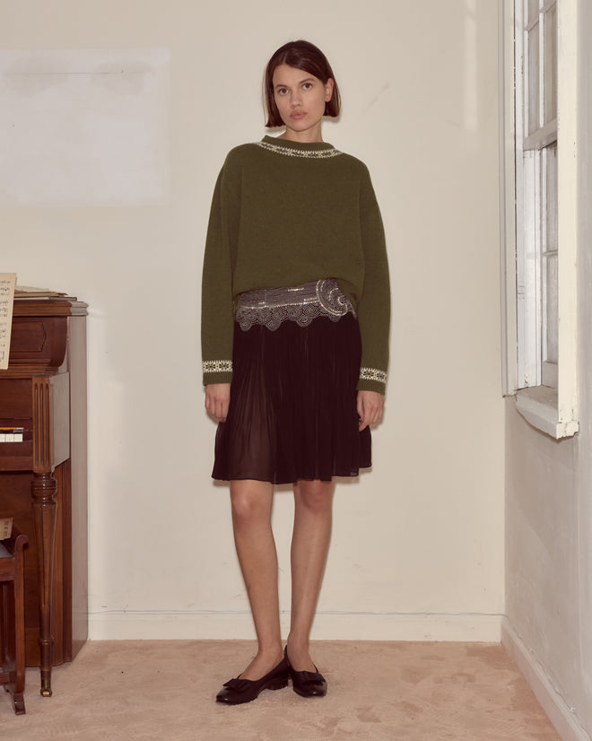 A woman wearing an olive green sweater, the Ensemble Skirt in black silk georgette with beading, and black flats stands indoors by a piano and window, evoking a 1930s reproduction style.