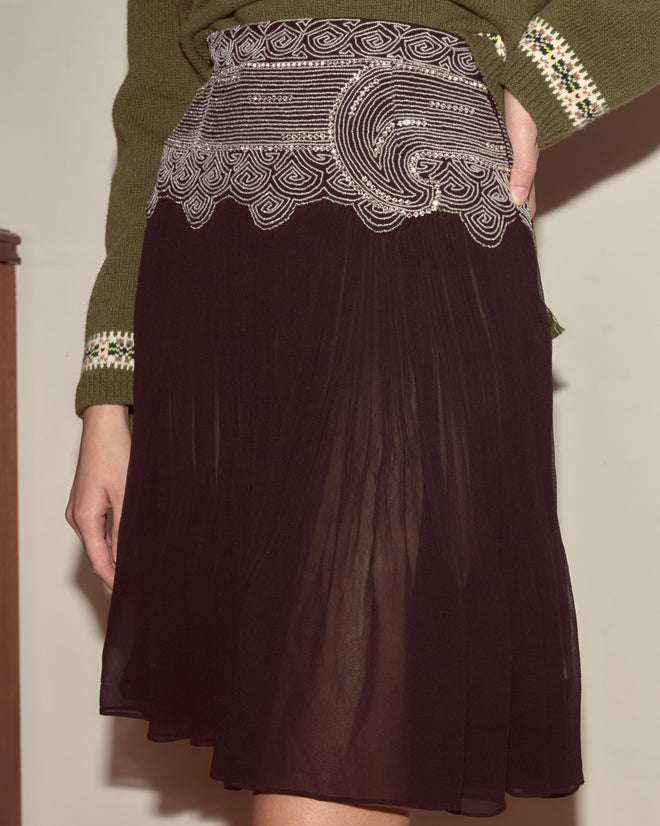 A person wearing the Ensemble Skirt—black with 1930s-inspired beading and white embroidery—paired with a green sweater featuring patterned cuffs.