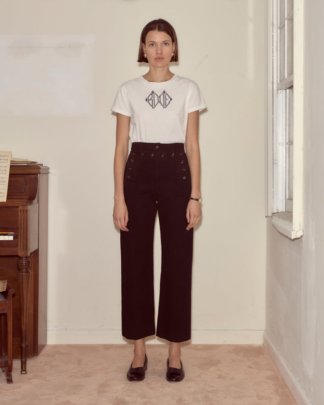 A woman stands indoors by a piano and window, wearing Denim Sailor Trousers - Black and a white graphic t-shirt.
