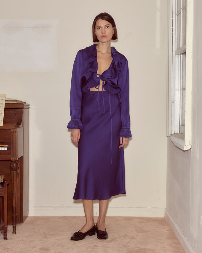 A woman in a deep blue ruffled blouse and the Dream Souvenir Skirt stands indoors near a piano and window.