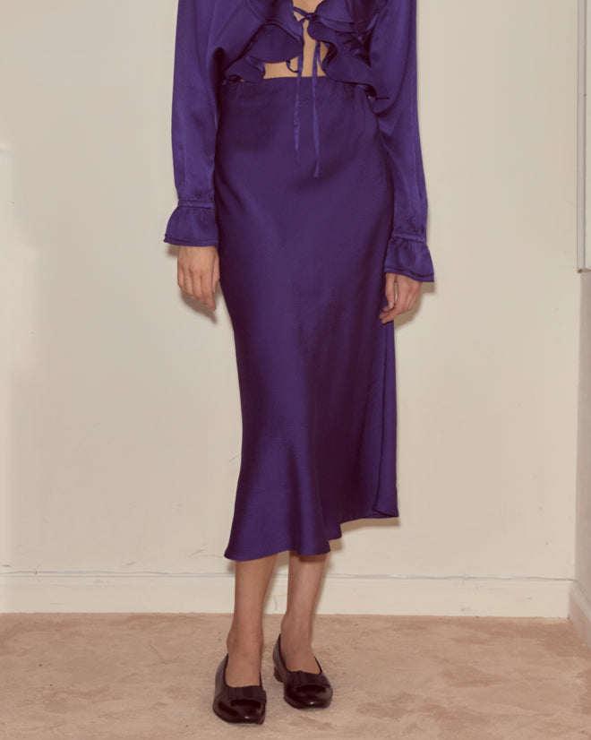 A person wearing the Dream Souvenir Skirt in silky purple-blue with a matching ruffled top and black shoes, standing indoors.