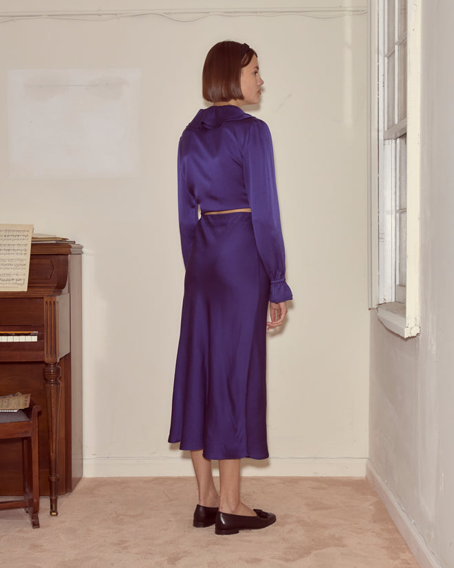 A woman wearing a long-sleeved purple dress featuring the Dream Souvenir Skirt stands facing a window in a simple room with a piano.