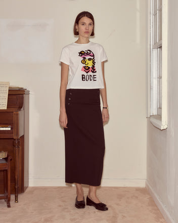 A woman in the Visage Tee and a long black skirt stands indoors by a piano and window on a beige carpet, her outfit echoing abstract colorblocking inspired by modernist art.