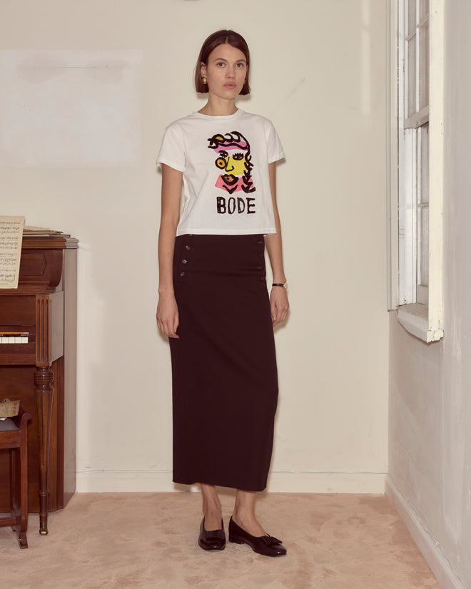 A woman in the Visage Tee and a long black skirt stands indoors by a piano and window on a beige carpet, her outfit echoing abstract colorblocking inspired by modernist art.