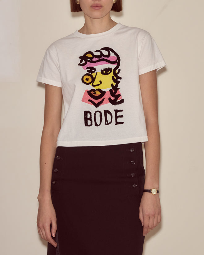 A woman wears the Visage Tee, a white BODE t-shirt featuring an abstract face inspired by Modernist art, paired with a black skirt.