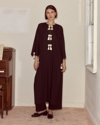 A woman wearing the Shadow Kaftan, a long black wool crepe dress with white tassels, stands indoors by a piano and a window.