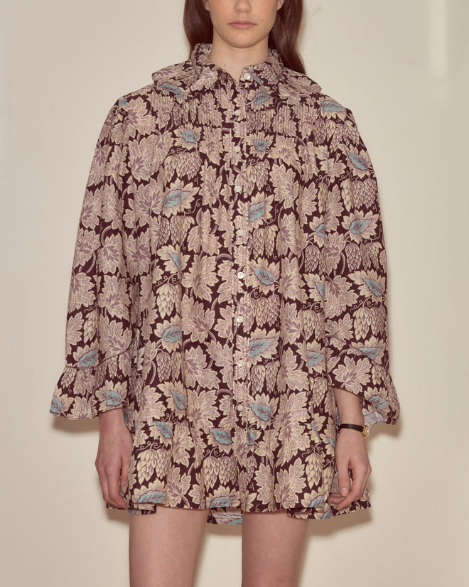 A woman stands against a plain wall wearing the Wisteria Dress, a loose brown and beige floral-patterned piece with blue accents, inspired by vintage nightgown styles.