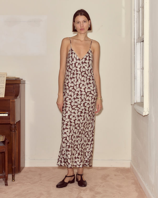 A woman in a Paper Birch Slip Dress stands indoors by a piano and window, gazing at the camera.