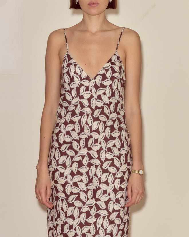 Woman wearing the Paper Birch Slip Dress, a sleeveless slip dress with a white leaf-stamp pattern on a dark background, standing against a plain wall.