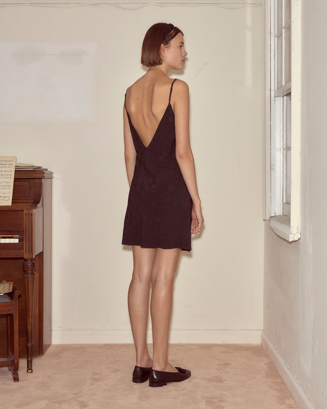A woman in the Moonflower Jacquard Short Slip, featuring a black silk design and low back, stands by a window in a softly lit room, facing away.