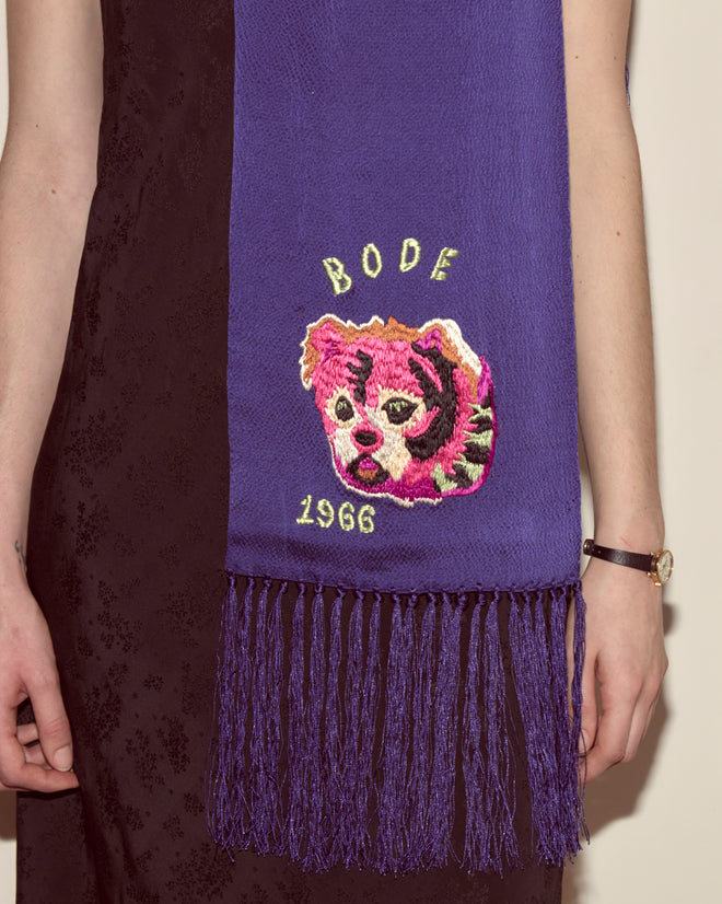 Person wearing the Dream Souvenir Scarf by BODE—purple textured silk with a pink tiger face, “1966,” and decorative fringe at the ends.