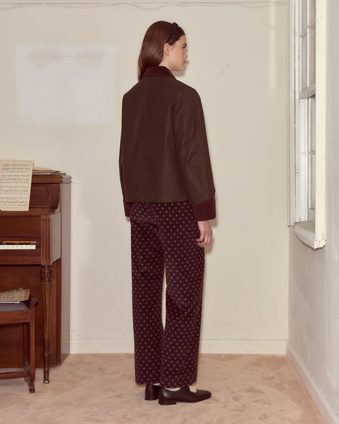 A person stands indoors facing away, near a piano with sheet music, wearing patterned pants and the Waxed Craft Jacket - Brown featuring subtle corduroy details.