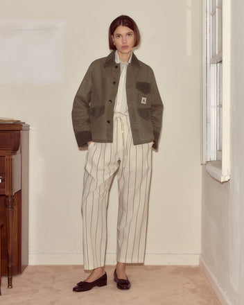 A woman stands indoors wearing the Waxed Craft Jacket - Light Green with corduroy trim over a striped cream shirt, cream pants, and black flats.
