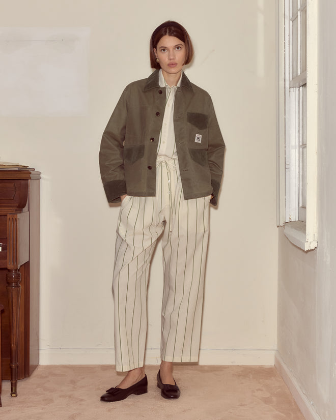 A woman stands indoors wearing the Waxed Craft Jacket - Light Green with corduroy trim over a striped cream shirt, cream pants, and black flats.