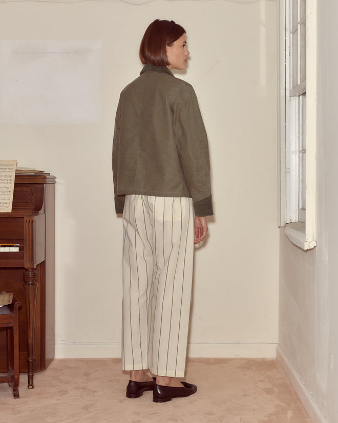 Woman in striped pants, Waxed Craft Jacket - Light Green, and black shoes stands in a room near a window, facing away.