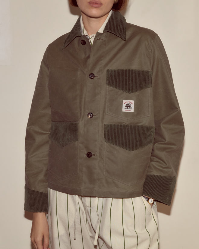A person wears the Waxed Craft Jacket - Light Green with corduroy trim and striped pants, standing against a beige wall.