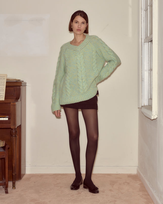 Woman in a cozy Eiderdown Sweater - Seafoam, black shorts, and black tights stands indoors by a piano and a window.