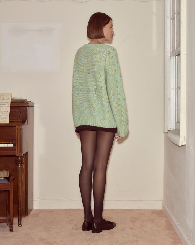 A woman in the Eiderdown Sweater - Seafoam, paired with black shorts and tights, stands by a window in a cozy room, facing away.