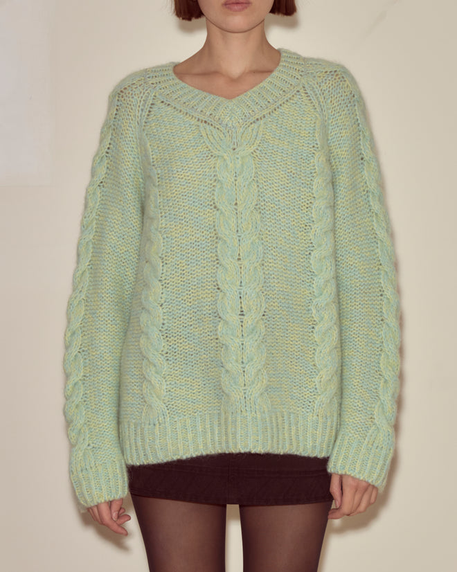 A person wearing the Eiderdown Sweater in Seafoam, a light green chunky cable-knit baby alpaca piece, paired with a black skirt, stands against a plain background.