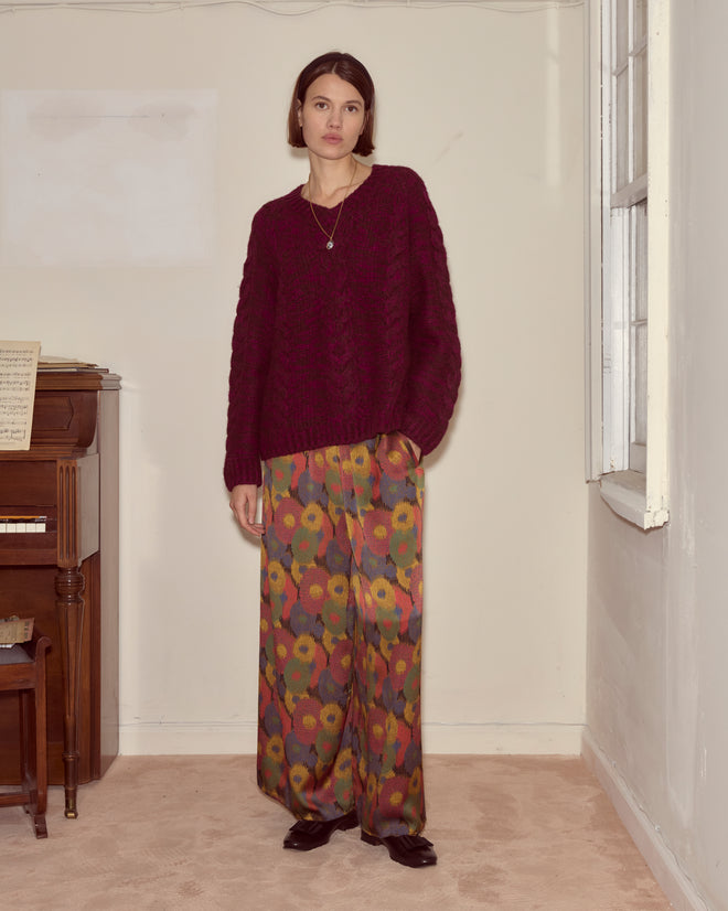 A woman wearing the Eiderdown Sweater - Maroon and colorful wide-leg pants stands indoors beside a piano and a window.
