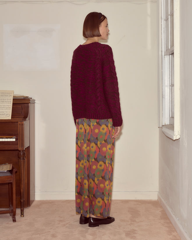 A woman stands sideways in a room, wearing the Eiderdown Sweater in maroon and colorful floral wide-leg pants.