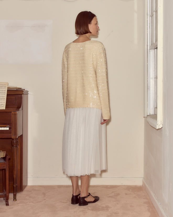 A woman in a Sequin Spotlight Swing Cardigan - Ivory and a white skirt stands indoors, facing a corner near a window and piano.
